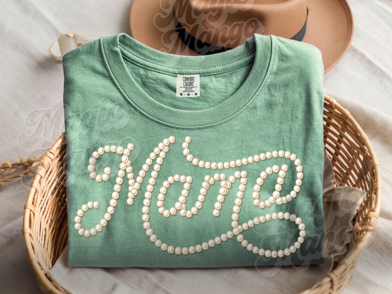 May include: A sage green t-shirt with the word "Mama" spelled out in a cursive font, using small, round, white embellishments. The shirt is folded and placed in a woven basket, with a brown hat visible in the background.