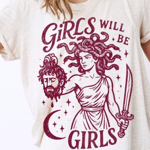 May include: Cream-colored t-shirt with a maroon graphic of Medusa holding a severed head and a sword. The text "GIRLS WILL BE GIRLS" is above and below the illustration. The shirt is a casual, short-sleeved style.
