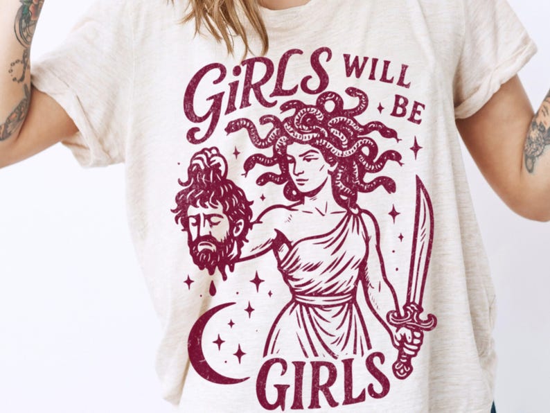 May include: Cream-colored t-shirt with a maroon graphic of Medusa holding a severed head and a sword. The text "GIRLS WILL BE GIRLS" is above and below the illustration. The shirt is a casual, short-sleeved style.
