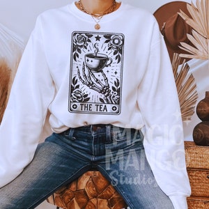 May include: White sweatshirt featuring a black and white tarot card design. The graphic depicts a skeletal hand holding a teacup, surrounded by roses and the text "THE TEA". The sweatshirt is paired with blue jeans.