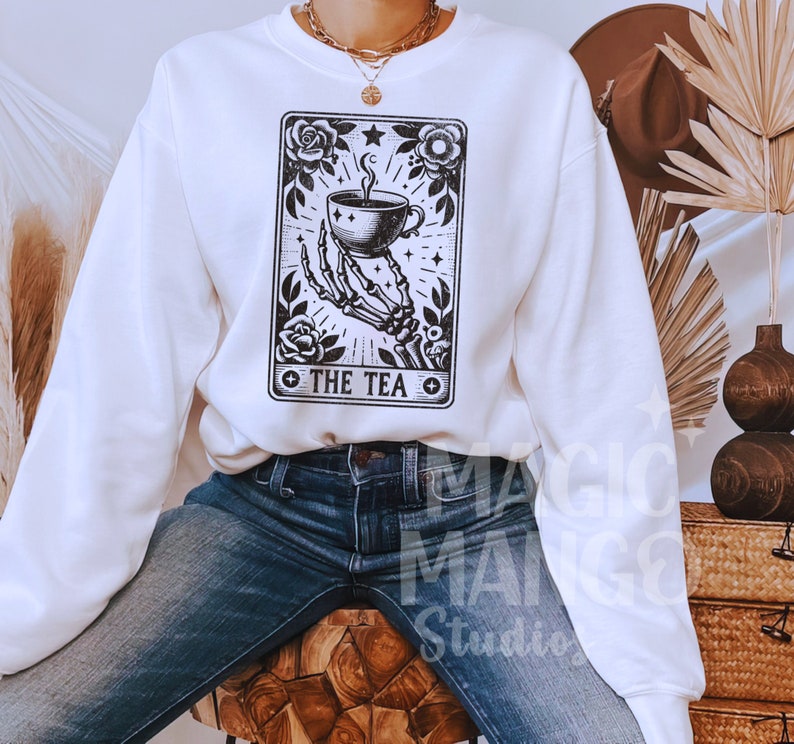 May include: White sweatshirt featuring a black and white tarot card design. The graphic depicts a skeletal hand holding a teacup, surrounded by roses and the text "THE TEA". The sweatshirt is paired with blue jeans.