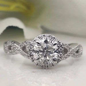 May include: A diamond engagement ring with a round center stone and a halo setting. The ring features a twisted band design and is set against a blurred white background with a white flower.