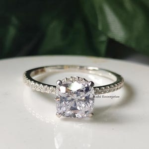 May include: A silver ring with a large, square-cut, clear gemstone as the centerpiece. The band is encrusted with smaller, clear stones. The ring is set against a soft, white background. The text "Ruhi Enterprise" is visible.