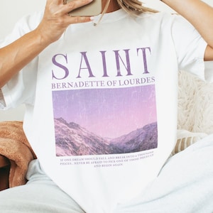 May include: White t-shirt with a purple graphic print of a mountain range and the text "SAINT BERNADETTE OF LOURDES". The text "If one dream should fall and break into a thousand pieces, never be afraid to pick up one of those pieces and begin again." is printed below the graphic.