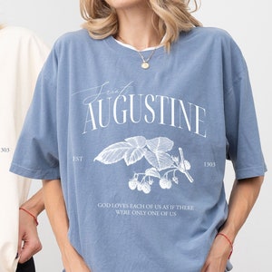 May include: Two women's t-shirts with a vintage-inspired design. The shirts are in ivory and blue with a white graphic of a blackberry branch and the text "EST AUGUSTINE 1303" and "GOD LOVES EACH OF US AS IF THERE WERE ONLY ONE OF US".