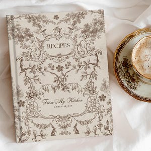 Puede incluir: Un libro de recetas con la palabra "RECIPES" en la portada y el texto "From My Kitchen". Junto a él, una taza de café sobre un platillo con borde dorado. Los artículos están sobre una sábana blanca.