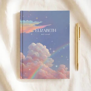 Puede incluir: Un cuaderno personalizado con un diseño de cielo de ensueño que presenta nubes, un arcoíris y estrellas. La portada muestra el nombre "ELIZABETH" y el año "EST 2016" en blanco. Un bolígrafo dorado descansa junto al cuaderno.