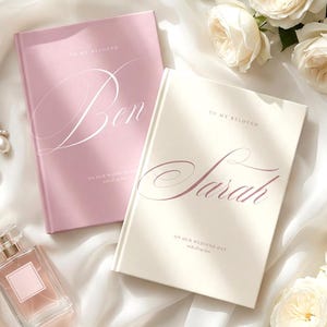 Op de afbeelding: Twee trouwdagboeken, één roze en één crème, met elegante scriptnamen en de woorden "To My Beloved" en "On Our Wedding Day." De afbeelding bevat witte rozen, een parfumflesje en pareloorbellen.