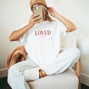 May include: White t-shirt with the word "LOVED" in red lettering and the text "ROMANS 5:8" in smaller red lettering.