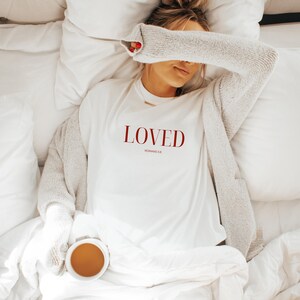 May include: White t-shirt with the word "LOVED" in red lettering and the text "ROMANS 5:8" in smaller red lettering below. The shirt is worn by a person lying in bed.