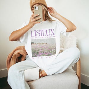 May include: White t-shirt with the word "LISIEUX" in purple, along with the phrases "DO ALL THAT YOU DO" and "WITH LOVE". The shirt features a landscape image of a field of purple flowers. A brown beanie and light gray sweatpants are also visible.