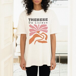 以下が含まれることがあります： ピンクとオレンジの色合いのスタイリッシュな花が描かれた白いTシャツ。Tシャツには「Therese De Lisieux」の文字がプリントされています。