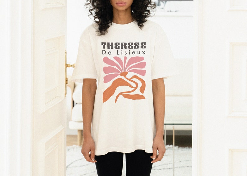 以下が含まれることがあります： ピンクとオレンジの色合いのスタイリッシュな花が描かれた白いTシャツ。Tシャツには「Therese De Lisieux」の文字がプリントされています。