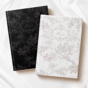 May include: Two hardcover journals with a floral pattern. The journal on the left is black with a silver floral pattern and the word "Vows" in a silver oval. The journal on the right is white with a gray floral pattern and the word "Vows" in a gray oval.