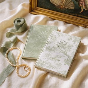 May include: Two hardcover vow books with sage green and white toile de jouy patterns. One book is titled His Vows and the other Her Vows, both featuring the names Noah & Emily and a date. A string of pearls, a gold ring, and sage green ribbon.