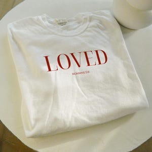 May include: White t-shirt with the word "LOVED" in red lettering and the text "ROMANS 5:8" in smaller black lettering.