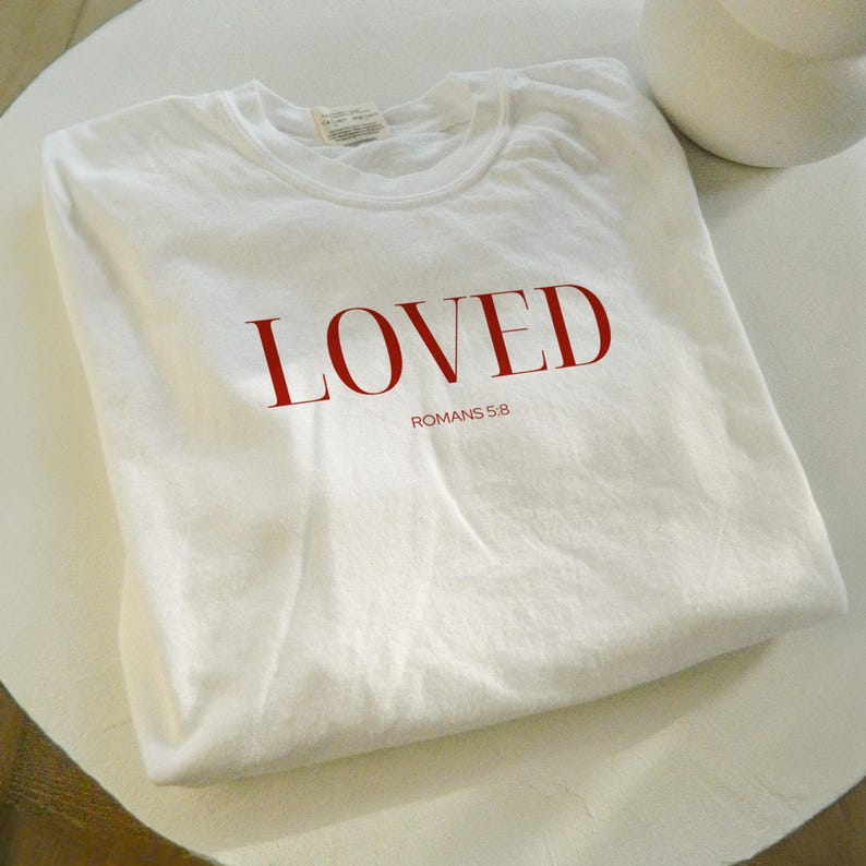 May include: White t-shirt with the word "LOVED" in red lettering and the text "ROMANS 5:8" in smaller black lettering.