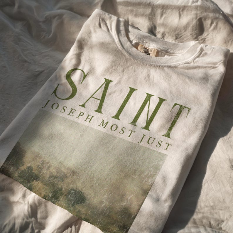 Może przedstawiać: Biała koszulka z zielonym nadrukiem graficznym, na kt&oacute;rym widnieje napis "SAINT JOSEPH MOST JUST", a pod nim znajduje się rozmyte zielone zdjęcie krajobrazu.