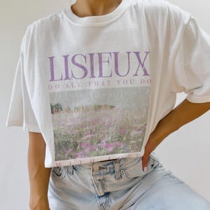 May include: White t-shirt with the word "LISIEUX" in purple block letters above a landscape image of a field of purple flowers. The text "DO ALL THAT YOU DO" is below the word "LISIEUX". The words "WITH LOVE" are at the bottom of the image.
