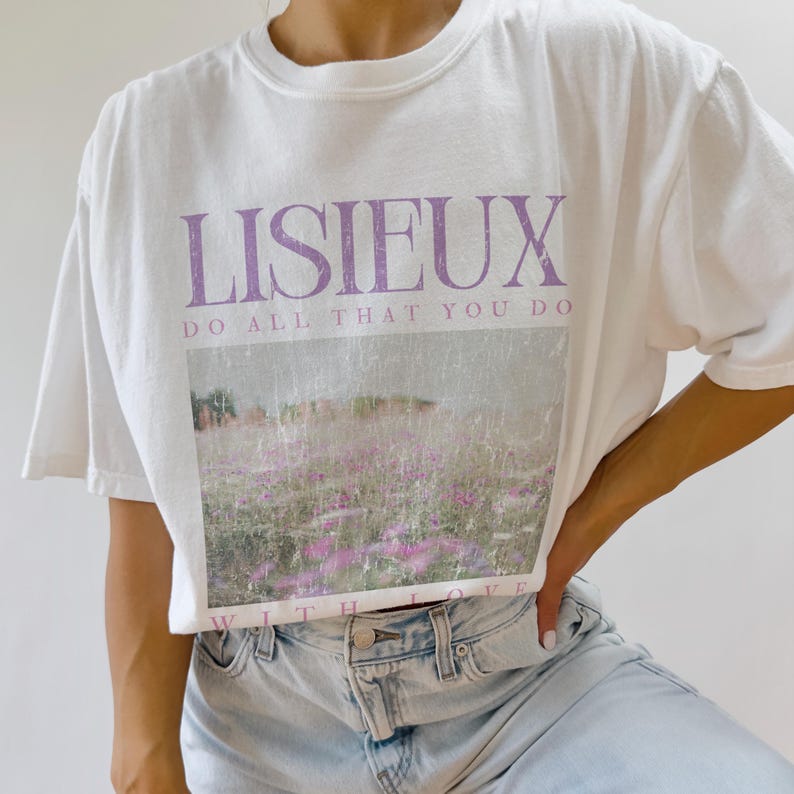May include: White t-shirt with the word "LISIEUX" in purple block letters above a landscape image of a field of purple flowers. The text "DO ALL THAT YOU DO" is below the word "LISIEUX". The words "WITH LOVE" are at the bottom of the image.