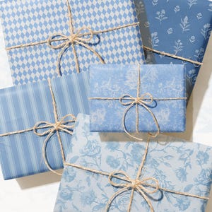 May include: An assortment of wrapped gifts in various shades of blue, tied with twine bows. The wrapping paper features patterns like diamonds, stripes, and floral designs. The gifts are arranged on a white surface.