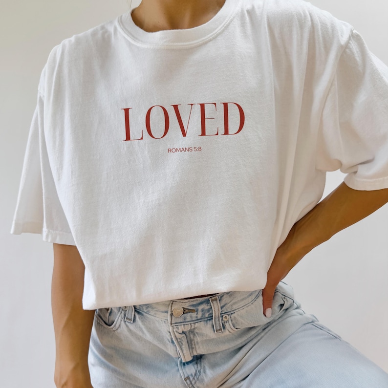 May include: White t-shirt with the word "LOVED" in red lettering and the text "ROMANS 5:8" underneath.
