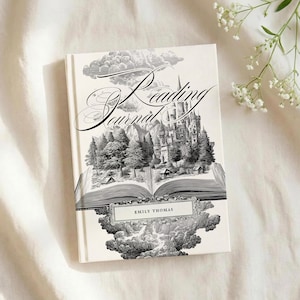Peut inclure: Un livre de journal de lecture &agrave; couverture rigide avec une illustration d&eacute;taill&eacute;e en noir et blanc d'un ch&acirc;teau, de montagnes et d'arbres. La couverture du livre porte le nom "Emily Thomas" et une &eacute;criture d&eacute;corative. Le livre est sur une surface blanche.