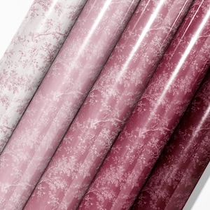 Puede incluir: Primer plano de varios rollos de papel de regalo decorativo en tonos rosados y rojos. Cada rollo presenta un delicado estampado floral en blanco, creando una estética suave y elegante. El acabado brillante realza los colores.