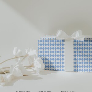 May include: A gift box wrapped in blue and white diamond patterned paper, tied with a white satin ribbon. White flowers are arranged to the left of the box. The words "BIRTHDAYS", "HOLIDAYS", and "WEDDINGS" are below the box.