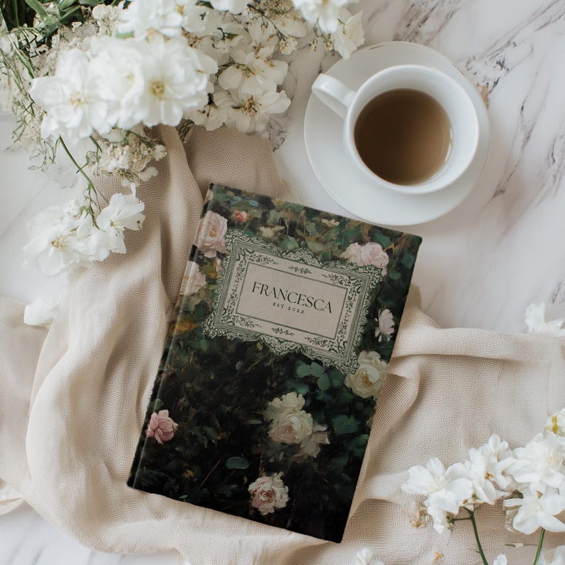 K&ouml;nnte beinhalten: Ein geschlossenes Hardcover-Journal mit einem floralen Design auf dem Umschlag. Der Umschlag zeigt einen dunkelgr&uuml;nen Hintergrund mit rosa und wei&szlig;en Rosen. Der Titel "FRANCESCA EST 2022" ist in einem goldenen Rahmen auf dem Umschlag gedruckt.