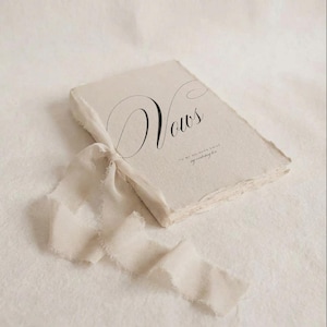 May include: A cream-colored vow book with the word "Vows" in elegant black script. The book is tied with a matching ribbon, and the pages have a deckled edge. The overall aesthetic is soft and romantic.