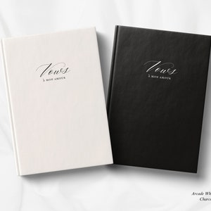 Puede incluir: Dos diarios de tapa dura con el texto "Vow's A Mon Amour" en una fuente cursiva. Un diario es blanco con texto negro y el otro es negro con texto blanco.