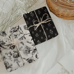 May include: Two wrapped gifts with floral patterns, one black and one white, tied with twine bows. The gifts rest on a white fabric surface, with a wicker basket and sprigs of white flowers in the background. The black gift has a small floral pattern.