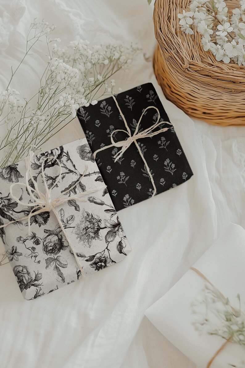 May include: Two wrapped gifts with floral patterns, one black and one white, tied with twine bows. The gifts rest on a white fabric surface, with a wicker basket and sprigs of white flowers in the background. The black gift has a small floral pattern.