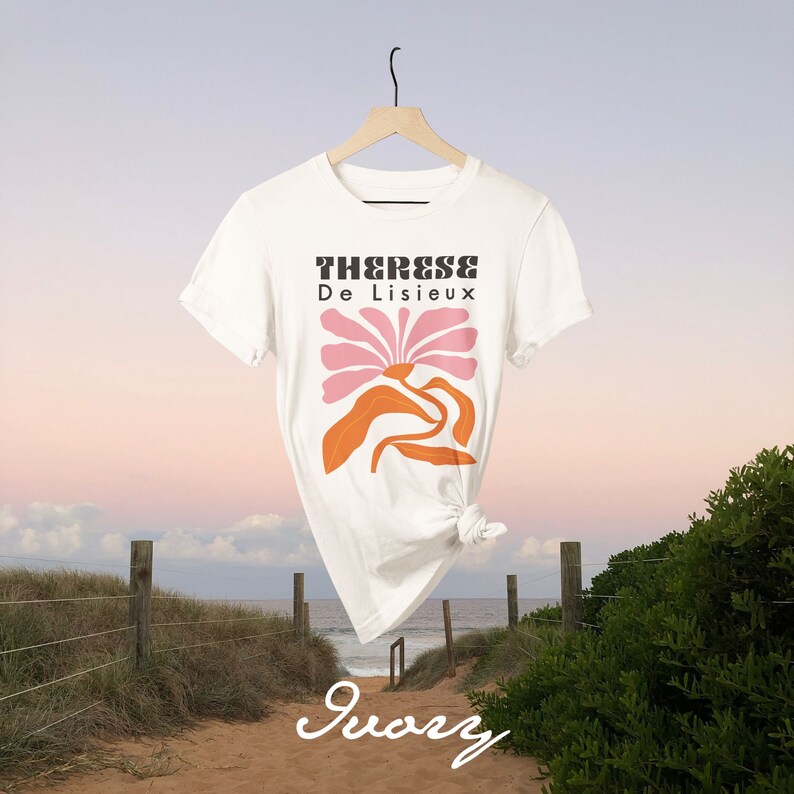 以下が含まれることがあります： 白いTシャツに、ピンクとオレンジの花のグラフィックと「Therese De Lisieux」という文字が前面にプリントされています。Tシャツは木製ハンガーにかけられ、ビーチの風景を背景にしています。