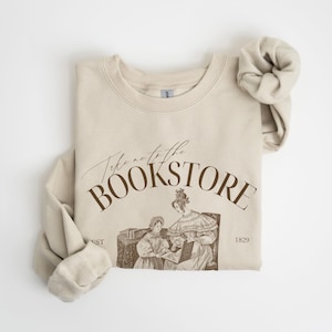May include: A beige sweatshirt with the text "Take me to the Bookstore" in brown lettering. The sweatshirt features a vintage illustration of two women reading a book.