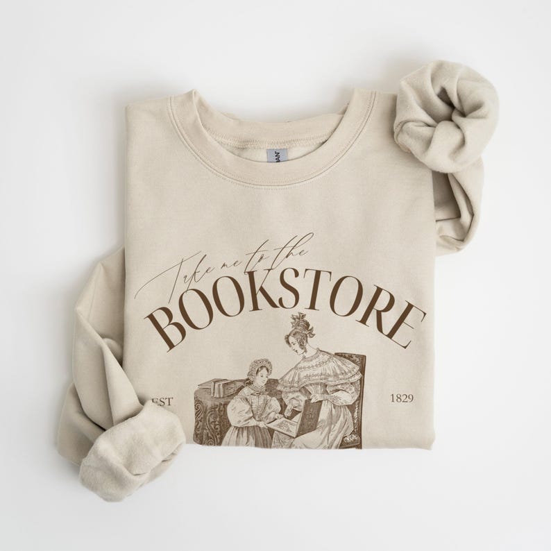 May include: A beige sweatshirt with the text "Take me to the Bookstore" in brown lettering. The sweatshirt features a vintage illustration of two women reading a book.