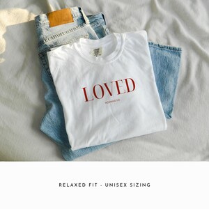 May include: White t-shirt with the word "LOVED" in red lettering and the text "ROMANS 5:8" underneath. The shirt is folded and laid on top of a pair of blue jeans.