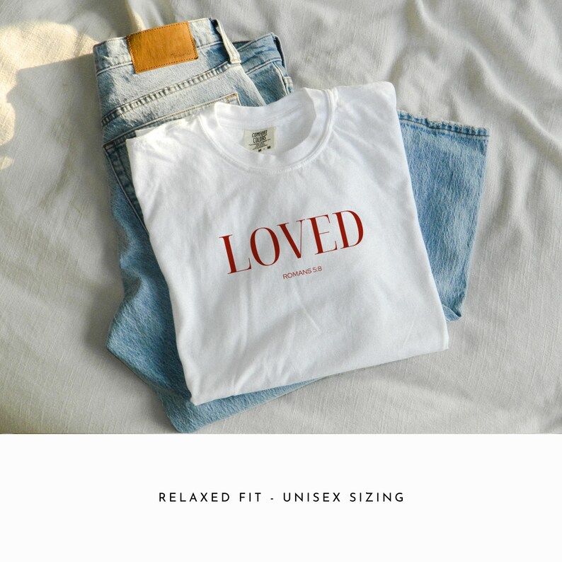 May include: White t-shirt with the word "LOVED" in red lettering and the text "ROMANS 5:8" underneath. The shirt is folded and laid on top of a pair of blue jeans.