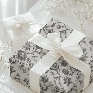 May include: Two gift boxes, one in the background and one in the foreground, are wrapped and tied with white ribbons. The larger box is covered in floral patterned paper in shades of gray and white. The smaller box is plain white.