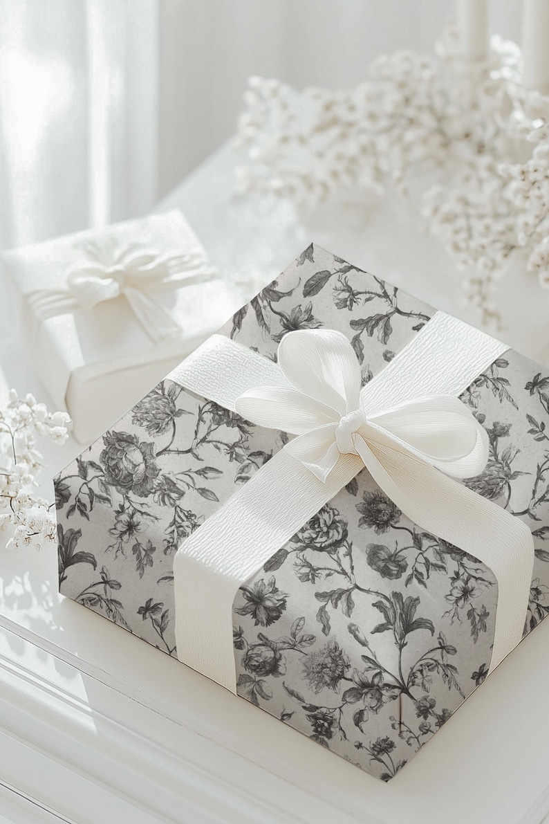 May include: Two gift boxes, one in the background and one in the foreground, are wrapped and tied with white ribbons. The larger box is covered in floral patterned paper in shades of gray and white. The smaller box is plain white.