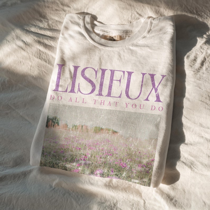 May include: A folded white t-shirt with the word "LISIEUX" in purple block letters. Below the word is the phrase "DO ALL THAT YOU DO". The shirt features a faded image of a field of purple flowers.