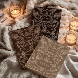 May include: Three recipe journals with floral designs and the word "Recipes" on the cover. The journals are in shades of brown and tan, with intricate white floral patterns. Lit candles in glass jars surround the journals, creating a cozy atmosphere.