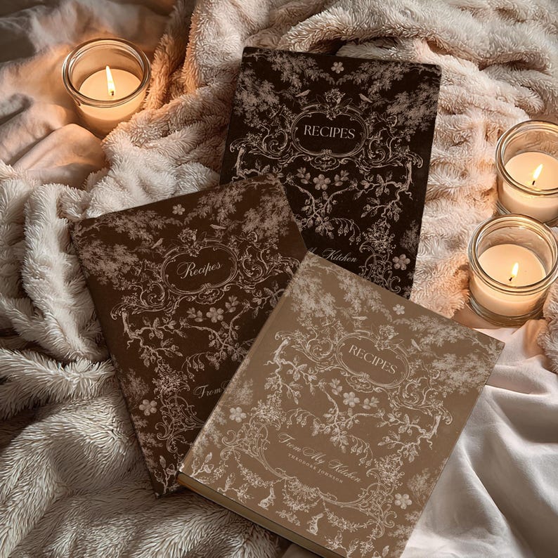 May include: Three recipe journals with floral designs and the word "Recipes" on the cover. The journals are in shades of brown and tan, with intricate white floral patterns. Lit candles in glass jars surround the journals, creating a cozy atmosphere.