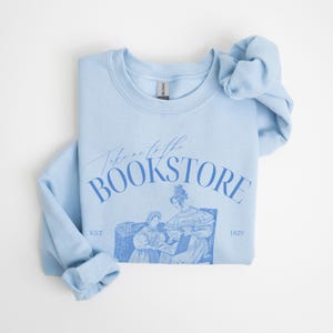 May include: Light blue sweatshirt with the text "Take me to the Bookstore" in blue lettering. The sweatshirt features a vintage illustration of two women in a bookstore.