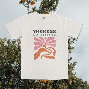 以下が含まれることがあります： ピンクとオレンジの花柄がプリントされた白いTシャツ。茶色の文字で「Therese De Lisieux」と書いてあります。