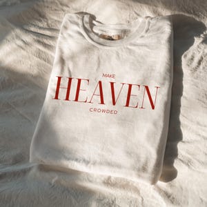 Op de afbeelding: Wit T-shirt met rode tekst die "MAKE HEAVEN CROWDED" luidt.