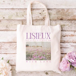 May include: Cream-colored tote bag with the word "LISIEUX" in purple script. Below the word is the phrase "DO ALL THAT YOU DO" and "WITH LOVE". The bag features a landscape image of a field of purple flowers.