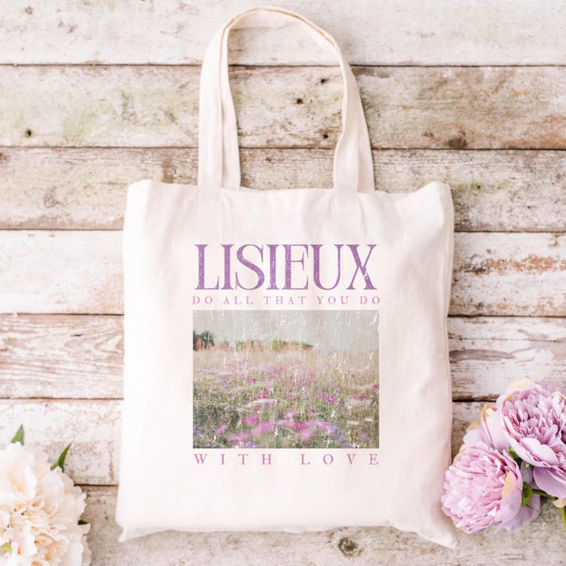 May include: Cream-colored tote bag with the word "LISIEUX" in purple script. Below the word is the phrase "DO ALL THAT YOU DO" and "WITH LOVE". The bag features a landscape image of a field of purple flowers.