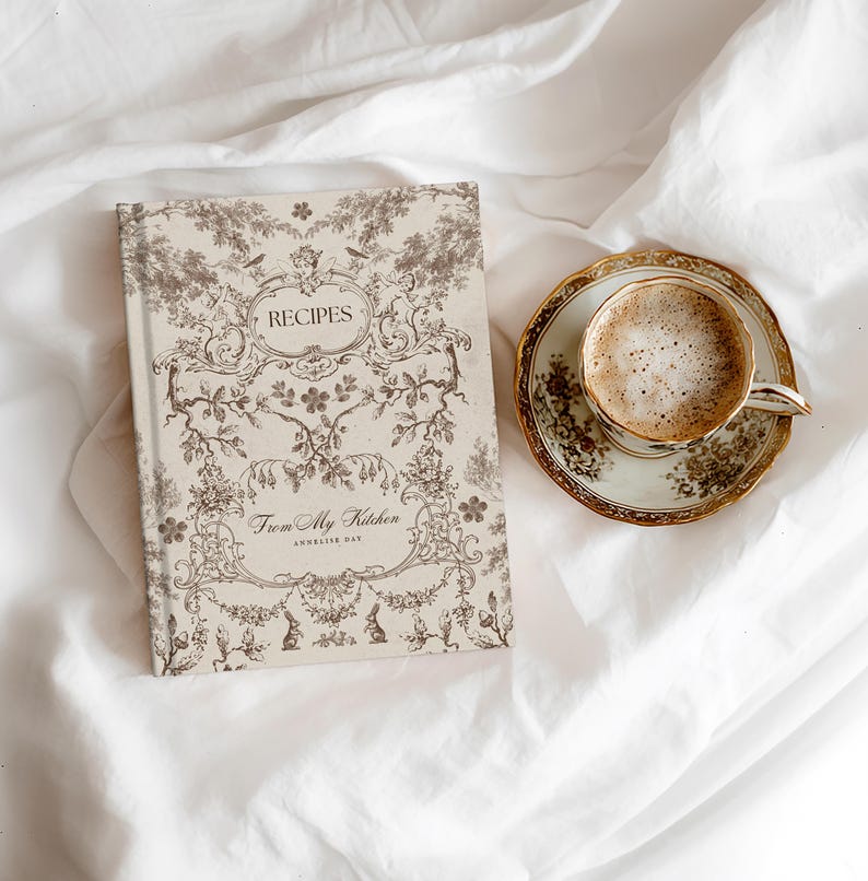 May include: A recipe book with the word "RECIPES" on the cover, along with the text "From My Kitchen". The book is next to a cup of coffee on a saucer with a gold rim. The items are on a white sheet.
